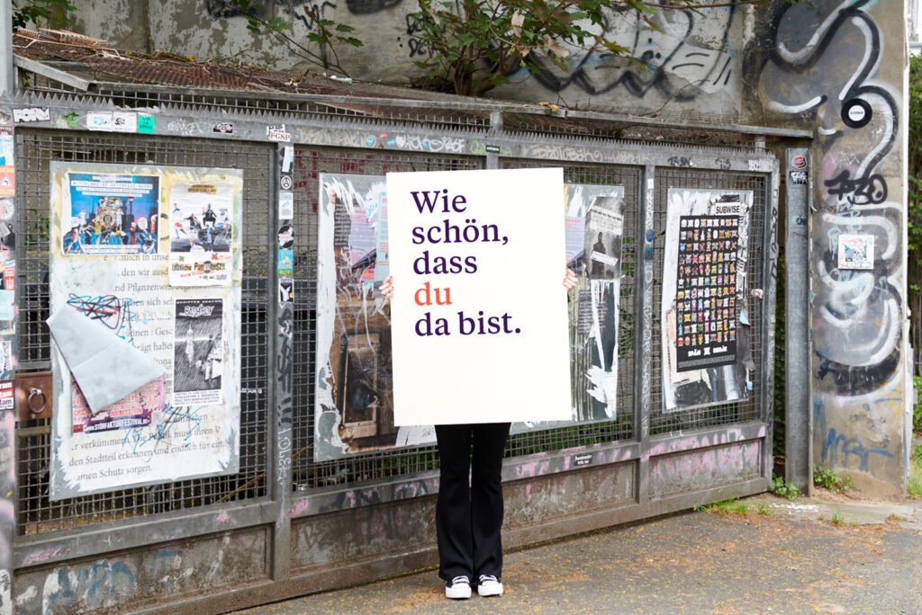 in Plakat mit der Aufschrift „Wie schön, dass du dabei bist.“ in einer urbanen Szene